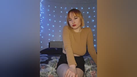 Video of a young, fair-skinned woman with blonde hair, wearing a tan long-sleeve top and black shorts, sitting on a bed with floral-patterned sheets, in a dimly lit room with blue fairy lights on a wall behind her.