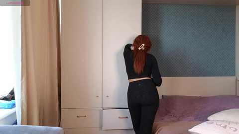 Video of a woman with long red hair in black workout clothes, adjusting a white wardrobe in a modern bedroom with patterned wallpaper, light-colored bed, and window.