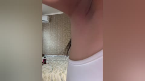 A close-up video of a person's arm and shoulder, wearing a white top, in a room with beige wallpaper, an air conditioner, and a bed with a white blanket.