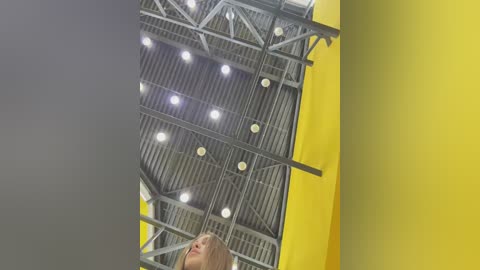 A video capturing a young girl with straight, light brown hair looking up at a yellow and grey industrial ceiling with exposed metal beams and numerous round lights.