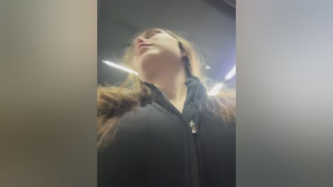 Video of a young woman with long brown hair, wearing a black jacket, standing indoors under fluorescent lighting. The background is blurred, emphasizing her upward gaze.
