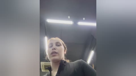 Video of a young Caucasian woman with light skin and brown hair, wearing a black jacket, seated in a dimly lit, modern interior with overhead fluorescent lights, possibly in a car or vehicle.
