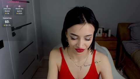 Video of a young woman with short black hair, wearing a red spaghetti-strap top, sitting on a bed in a dimly-lit bedroom.