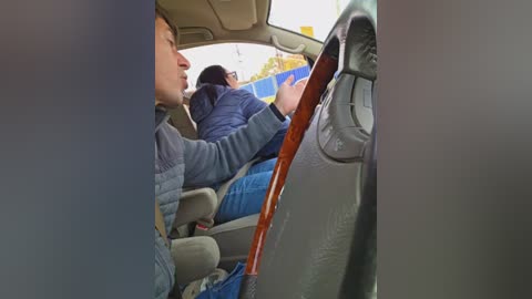 A video showing a man in a blue jacket sitting in a car, with another person behind him, and the car's interior visible, including a wooden steering wheel.