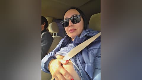 Video of a woman with light skin, wearing sunglasses and a blue jacket, driving a car with beige seats. She has a relaxed expression and is gripping the steering wheel.