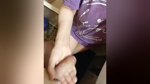 A video captures a woman's bare feet resting on a man's lap, wearing a purple T-shirt, in a dimly lit bathroom with a speckled floor and countertop.