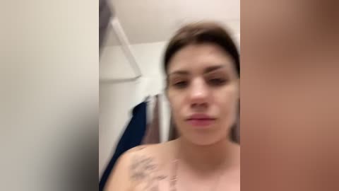 A blurred video of a young woman with light skin and brown hair, wearing a black top, standing in a bathroom. She has tattoos on her shoulder. A towel rack and a blue towel are visible in the background.