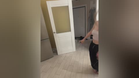 Video of a slender, light-skinned woman in black pajama pants and a white bra, opening a white door in a dimly lit, beige-walled hallway with wooden floors.