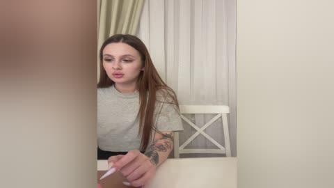 Video of a young woman with long brown hair, light skin, and tattoos, wearing a gray t-shirt, sitting at a table, looking at a phone, in a simple, well-lit room with white curtains and a white chair.