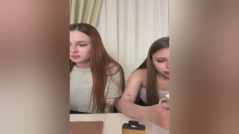 A video of two young women with long hair, one with straight brown hair and the other with straight blonde hair, sitting side by side in a bathroom.