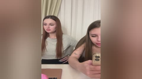 A video of a young woman with long brown hair, wearing a light grey top, taking a selfie in a bathroom mirror. She appears thoughtful.