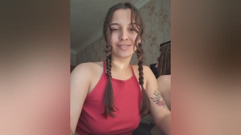 A video of a young Caucasian woman with light skin and brown hair in twin braids, wearing a red tank top, smiling. Background features patterned wallpaper and a blurred figure.