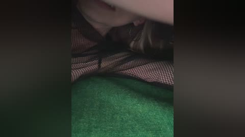 A video shows a close-up of a person's face, partially obscured by a black mesh mask, with green fabric beneath. The lighting is dim, creating a shadowy effect.