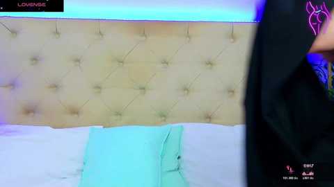 Video of a plush, beige, tufted headboard with a light blue pillow on a bed, partially visible. The background is a modern, minimalist room with blue lighting.