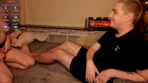 Video: A bald, muscular man in a black shirt and shorts, lying on a grey bed, appears to be masturbating while a smaller, nude, light-skinned woman with long hair, lying beside him, looks on. Background: Fireplace, wall, and digital game interface.