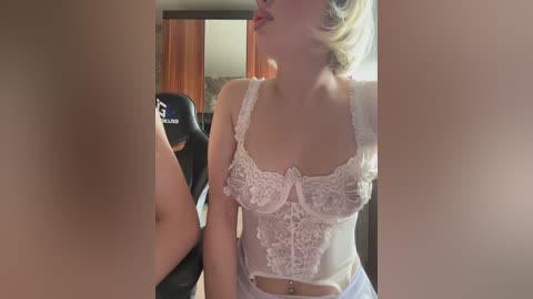 Video of a fair-skinned woman with blonde hair, wearing a lacy white bra and light blue skirt, standing in a dimly lit room with a gaming chair visible in the background.