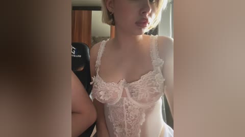 Video of a light-skinned woman with short blonde hair, wearing a delicate white lace lingerie bodysuit. She stands in a dimly lit room with a door and chair visible in the background.