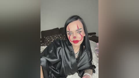 Video of a woman with pale skin, black hair, and red makeup, including red lips and face paint, wearing a black satin robe, lying on a bed with patterned pillows in a dimly lit room.
