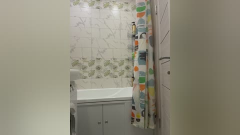 Video of a small bathroom with white tiled walls, a white sink and cabinet, and a colorful shower curtain featuring abstract patterns.