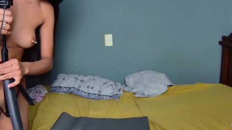 Video of a topless woman with medium breasts and light brown skin, standing by a bed with yellow sheets and patterned pillows, holding a microphone.