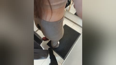 Video of a woman with long brown hair, wearing a beige sports bra and tight gray leggings, standing on a tiled floor with a black mat in a bathroom.