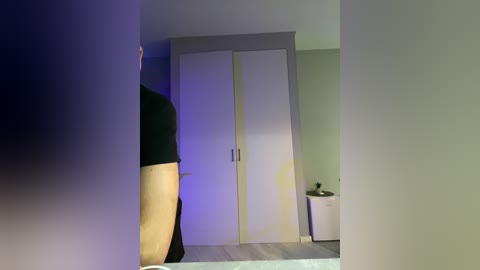 Video of a modern, minimalist bedroom featuring a white wardrobe, a dark-skinned person wearing a black shirt and beige pants, a white trash can, and a light grey wall.