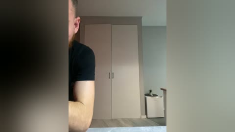 Video of a man with fair skin and red hair, wearing a black t-shirt, standing in a minimalist bedroom with beige walls, a built-in wardrobe, and a white sink.