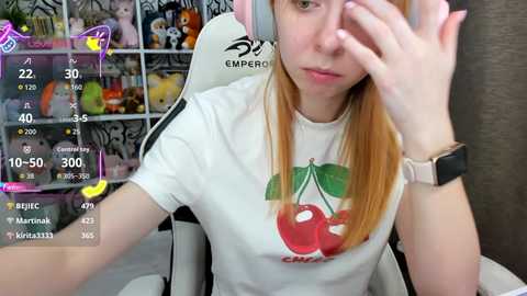 Video of a young woman with long blonde hair, wearing a white \"Cherry\" T-shirt, sitting in a white gaming chair, holding a smartphone, surrounded by colorful plush toys.