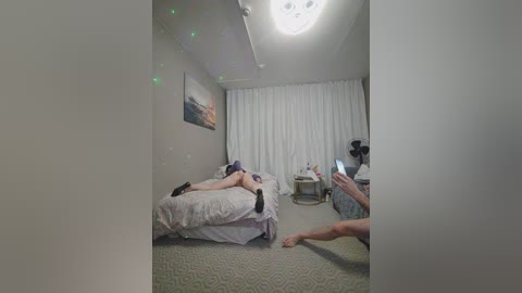 Video of a minimalist bedroom with a person lying on a bed, holding a phone, recording a selfie. The room features a large window with white curtains, a beige carpet, and a colorful painting on the wall.