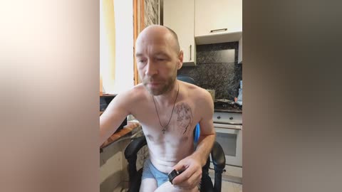 Video of a bald, muscular, light-skinned man with a beard, sitting shirtless in a kitchen. He wears blue boxers and holds a remote control, with a tattoo on his chest. The kitchen has white cabinets, a black granite countertop, and a blue chair.