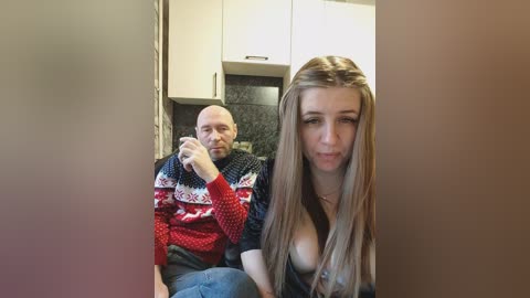 Video of a bald man in a red sweater, smoking, seated behind a young woman with long blonde hair, wearing a low-cut black top, in a modern kitchen with white cabinets and dark countertops.