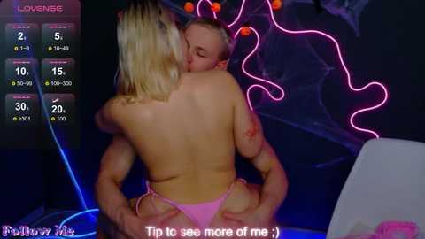 Video of a topless blonde woman with pink thong, sitting on a muscular man with a bloody face, in a dimly lit room with neon lights, with a game interface overlay showing \"Lover\" stats.