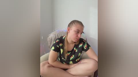 Video of a young woman with fair skin, braided hair, wearing a black t-shirt with green and yellow patterns, sitting cross-legged in a white room, appearing thoughtful.