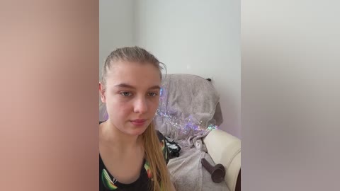 A video of a young Caucasian woman with light skin, straight blonde hair, and a neutral expression, wearing a colorful graphic tee, seated on a gray plush chair with a purple, glittery tulle skirt.