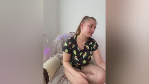 Video of a young woman with light skin and long blonde hair in a ponytail, sitting on a beige couch with a black shirt adorned with colorful cartoon fruit patterns.