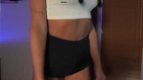 Video of a tan-skinned woman wearing a white crop top and black shorts, standing indoors with a wooden door and blinds in the background.