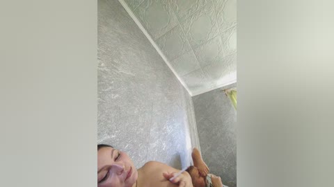 Video of a naked man lying on a textured, gray tiled floor, with his head resting on his hands, showing a calm, serene expression. The room has a white ceiling with a patterned design.