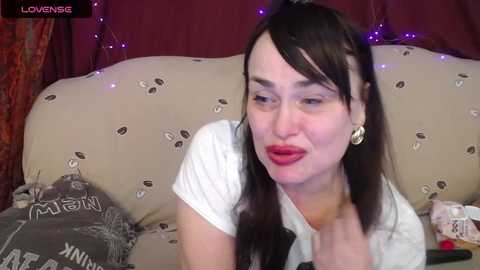 Video of a woman with straight black hair, wearing red lipstick, a white shirt, and a black tie, sitting on a beige couch with a gray pillow. Background includes a maroon curtain and purple string lights.