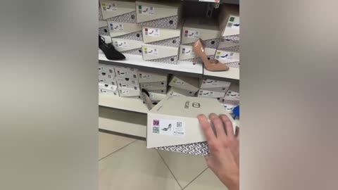 Video of a shoe display in a store, showing a hand holding a white shoebox with a barcode. The background features rows of shoe boxes and a pair of tan high heels.