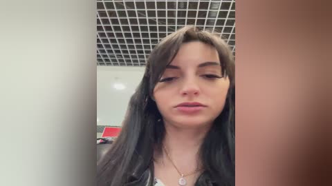 Video of a young woman with long black hair, fair skin, and dark eyes, wearing a black top, standing in a modern office with a grid ceiling.