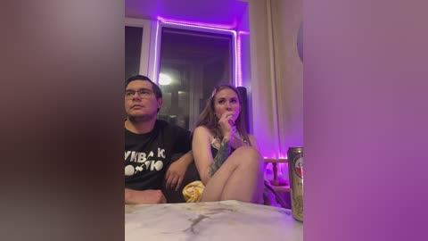 Video of a man in a black T-shirt and glasses, and a woman in a black bra and panties, sitting on a marble counter, both holding snacks under purple light.