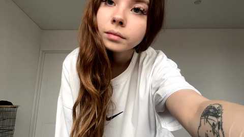 Video of a young Asian woman with long, wavy brown hair, wearing a white T-shirt, taking a selfie in a simple, light-colored room.