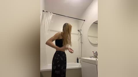 Video of a blonde woman in a black floral dress, standing in a small, beige bathroom with a white bathtub, showerhead, and sink.