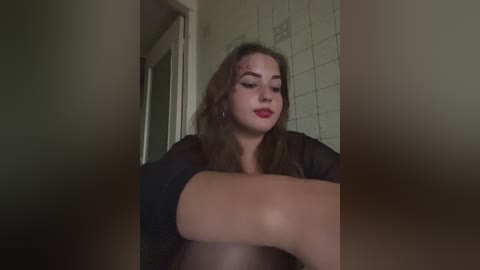 A video of a young woman with long, wavy brown hair, fair skin, and red lipstick, sitting in a dimly lit hallway with beige tiled walls. She wears a black top, looking contemplative.