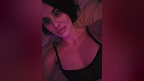 Video of a smiling woman with short black hair, wearing a black bra, lying on a bed under dim red lighting.