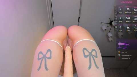 A video featuring a woman's spread legs, showcasing large, bow-shaped tattoos on her thighs, wearing sheer stockings. The background includes a medical equipment panel and a pink digital display.