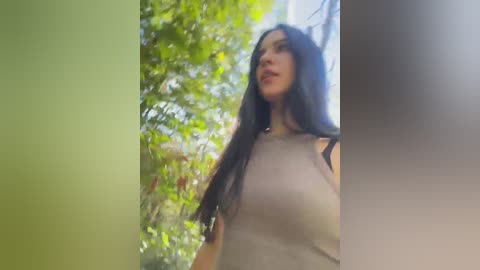 A video of a young woman with long black hair, wearing a beige tank top, standing in a green, sunlit garden.