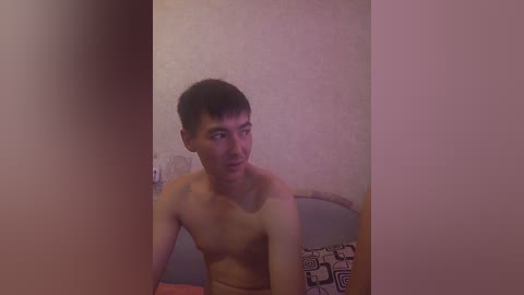 Video of a shirtless, slender Asian man with short dark hair, seated on a bed with patterned sheets, against a pink, textured wall, appearing slightly blurred.