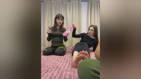 Video of two women, one in a green top and leggings, the other in a black top and leggings, on a pink bed, playing with pink socks, beige curtains in the background.