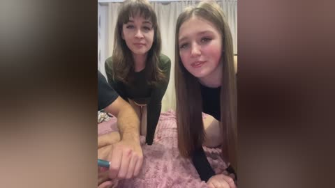 Video of two young women in a bedroom. One with long hair, wearing black, leans on bed. The other with short hair, wearing green, sits on bed.